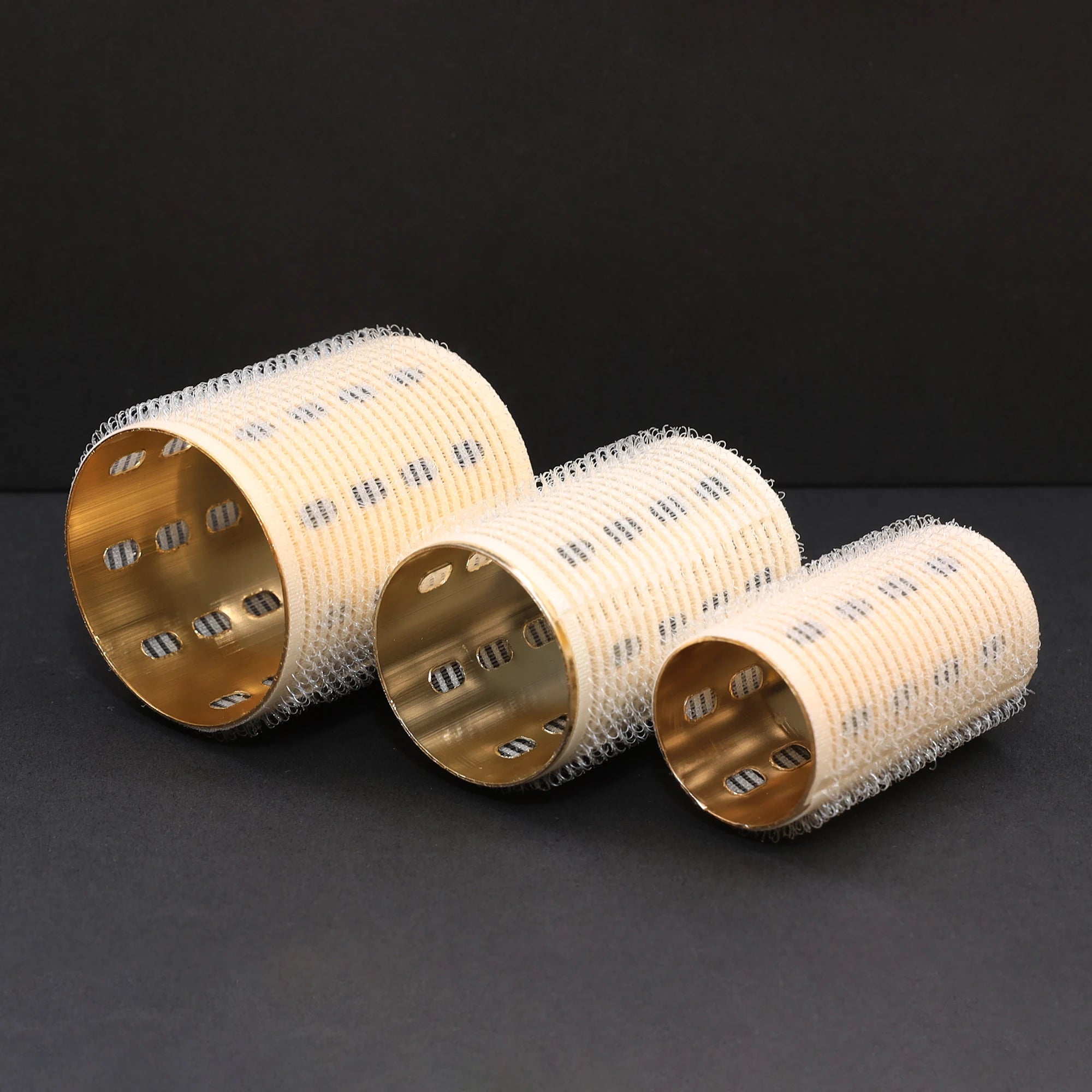 Three ceramic thermal hair rollers with gold and silver metallic finishes on a black background