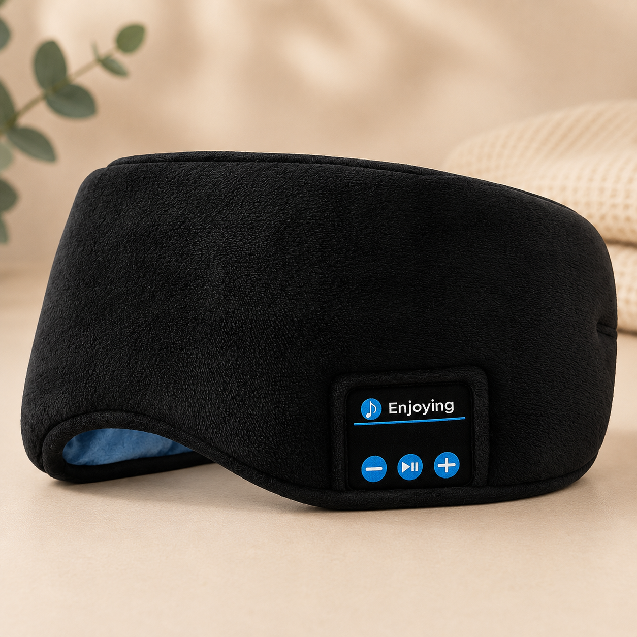 Black fleece bluetooth sleep eye mask with a built-in control panel displaying the 'Enjoying' brand logo and three blue buttons, set against a warm beige background with a blurred eucalyptus leaf.