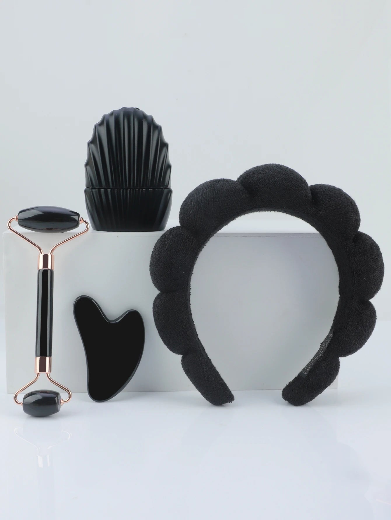 Set of black facial tools including a roller, gua sha tool, and headband on a light gray background