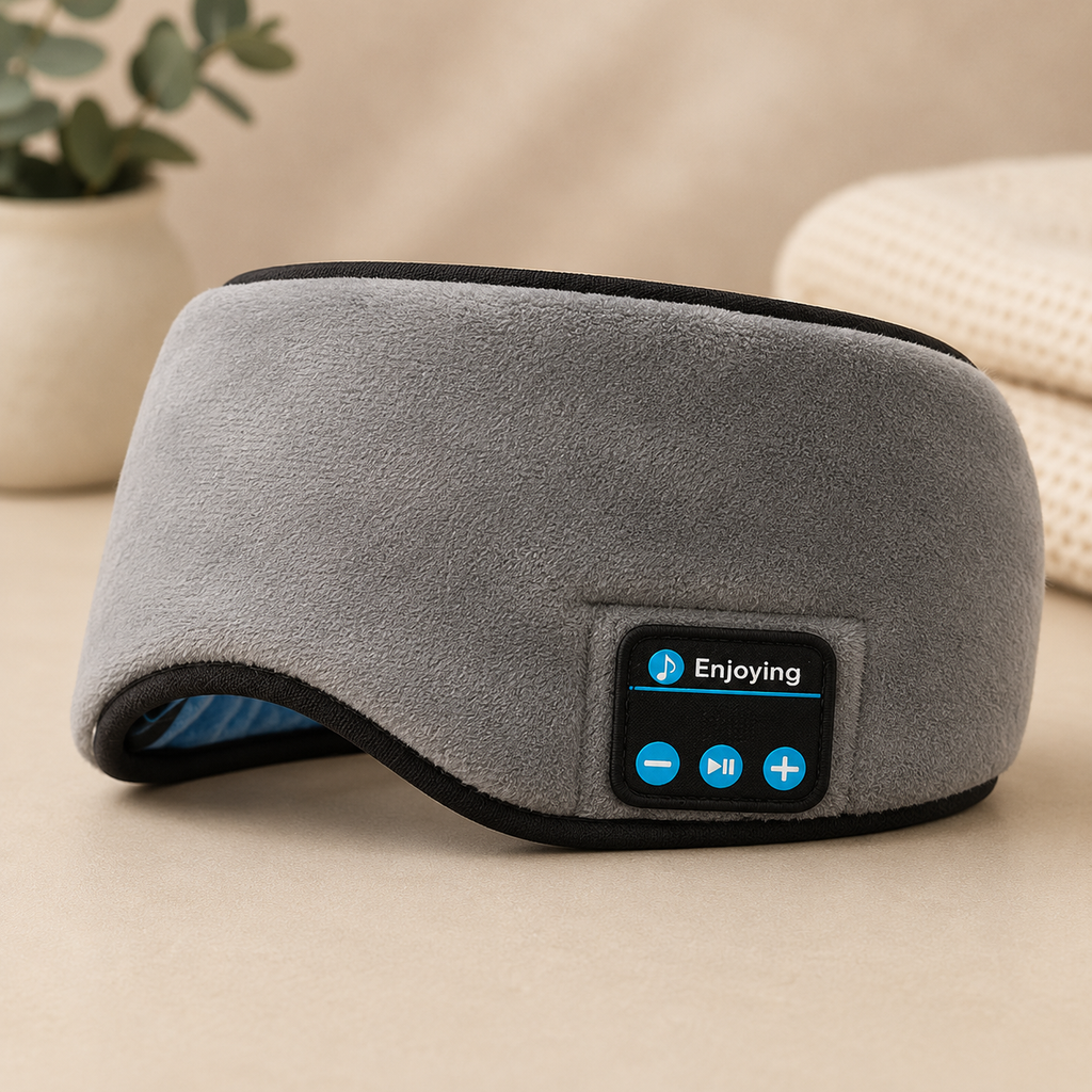 A gray fleece travel bluetooth sleep eye mask with black trim and a built-in control panel displaying blue icons and the word 'Enjoying'. The mask is shown resting on a beige surface with a blurred background containing a potted plant and a woven blanket.