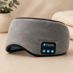 A gray fleece travel bluetooth sleep eye mask with black trim and a built-in control panel displaying blue icons and the word 'Enjoying'. The mask is shown resting on a beige surface with a blurred background containing a potted plant and a woven blanket.