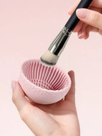 Hand holding a pink silicone brush cleaner with a makeup brush over it on a pink background
