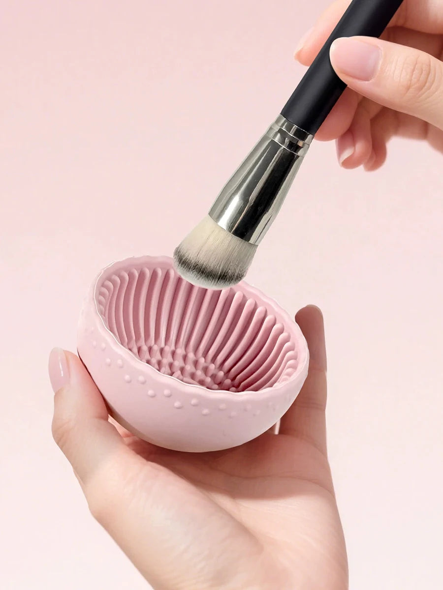 Hand holding a pink silicone brush cleaner with a makeup brush over it on a pink background