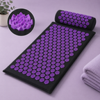Black acupressure mat with purple flower-shaped massage nodes, shown with a matching cylindrical pillow and a circular inset detail of the textured nodes. A folded light purple towel and a small plant are placed nearby on a pale lavender surface.