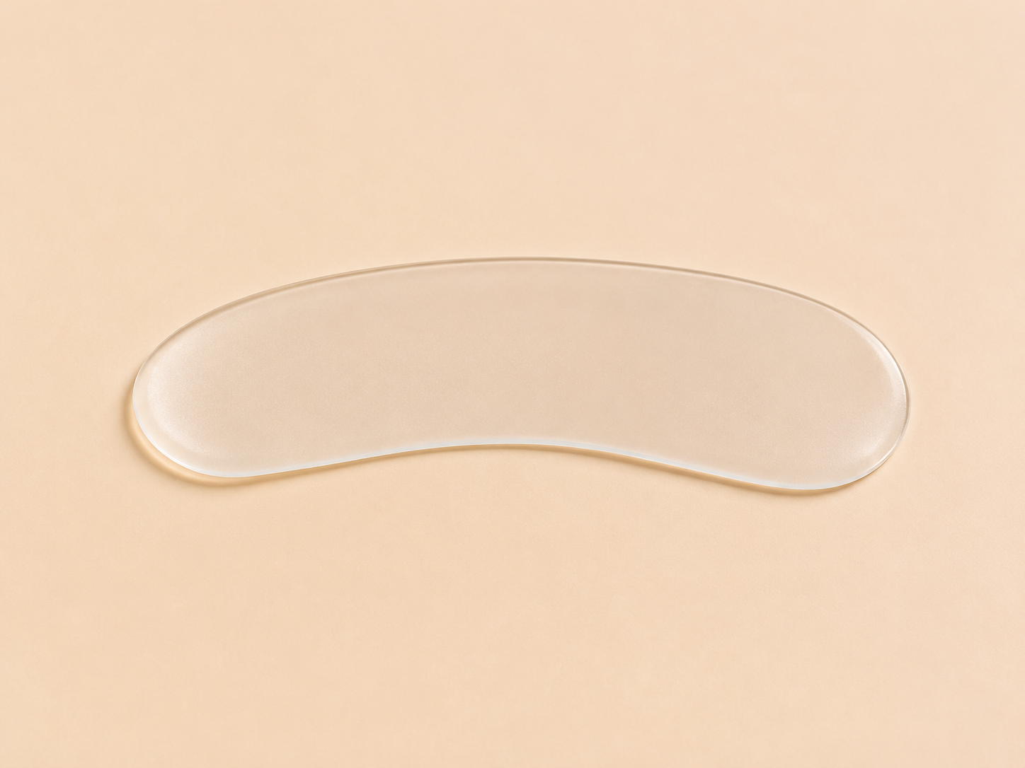 A clear, silicone curved gel pad or eye patch lying horizontally on a solid, warm beige background.