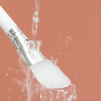 white face mask brush with water droplets against a peach-colored background