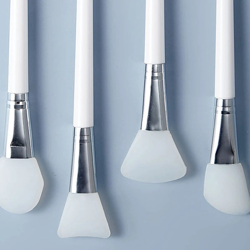 Four makeup face mask brushes with white handles and silicone bristle heads on a light blue background