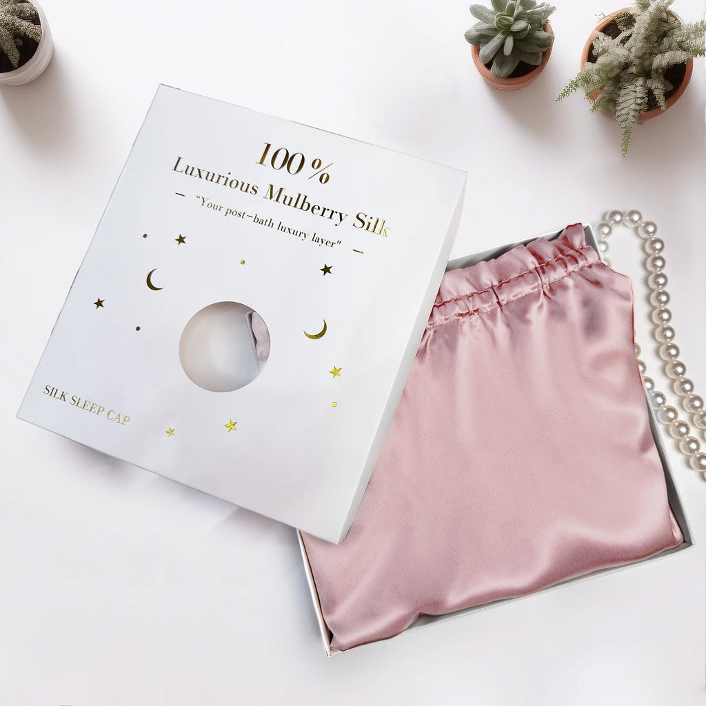 Pink  pure mulberry silk bonnet headscarf sleep cap with packaging on a white surface with plants in the background