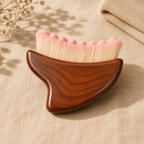 A wooden gua sha massage tool with soft pink and cream bristles, resting on a beige linen surface with dried flowers and a folded cloth in the background.